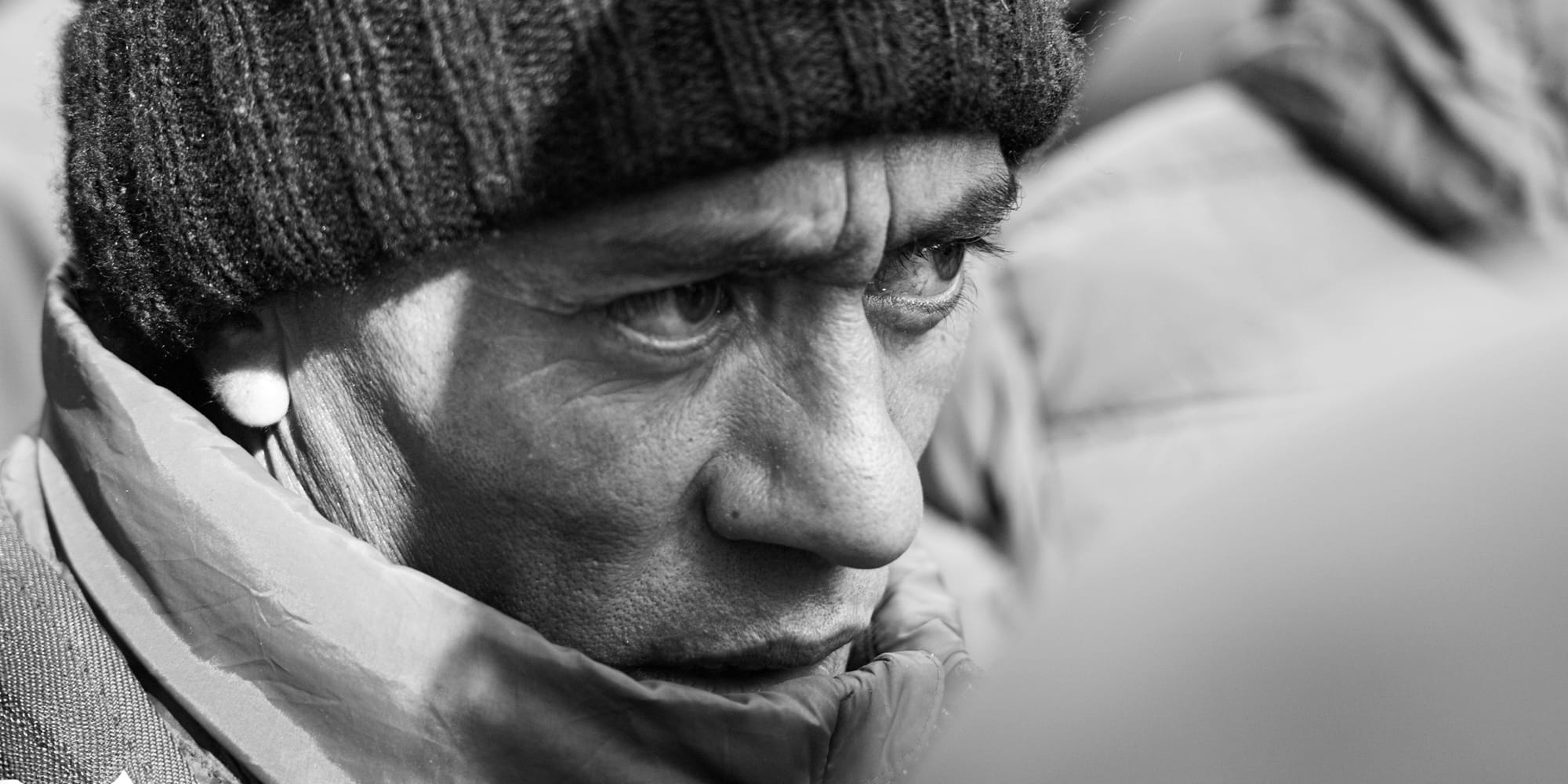 Black-and-white panoramic close-up portrait — the weathered face of an older man in a knit cap, brow furrowed, gazing into the middle distance amid the winter crowd.