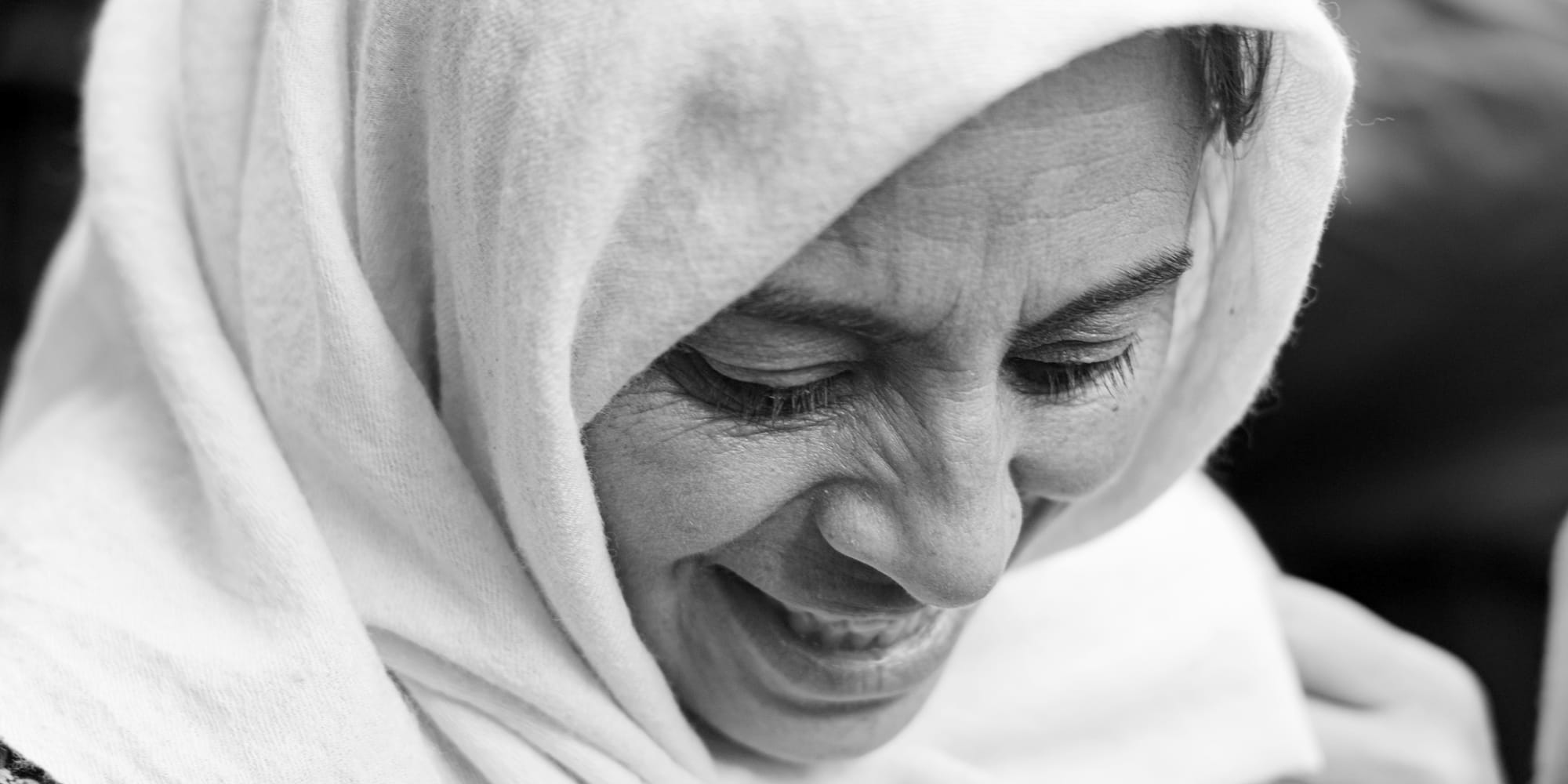 Black-and-white panoramic close-up portrait — an older woman in a pale headscarf creased with laughter, eyes shut, deep wrinkles catching the soft winter light.