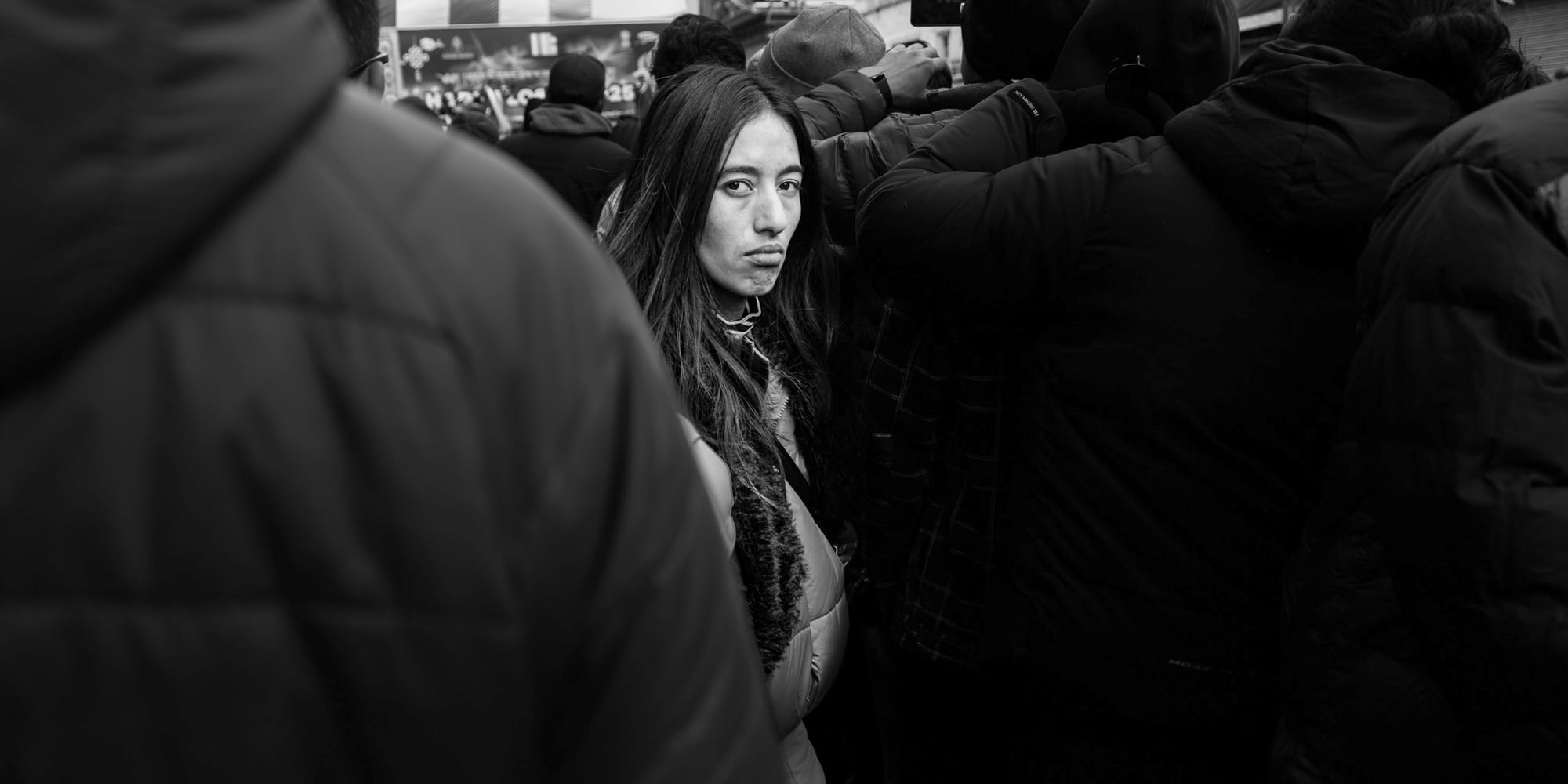 Black-and-white panoramic street portrait — a young woman with long dark hair holds a level, skeptical gaze, framed between two bundled strangers in a dense Losar festival crowd.