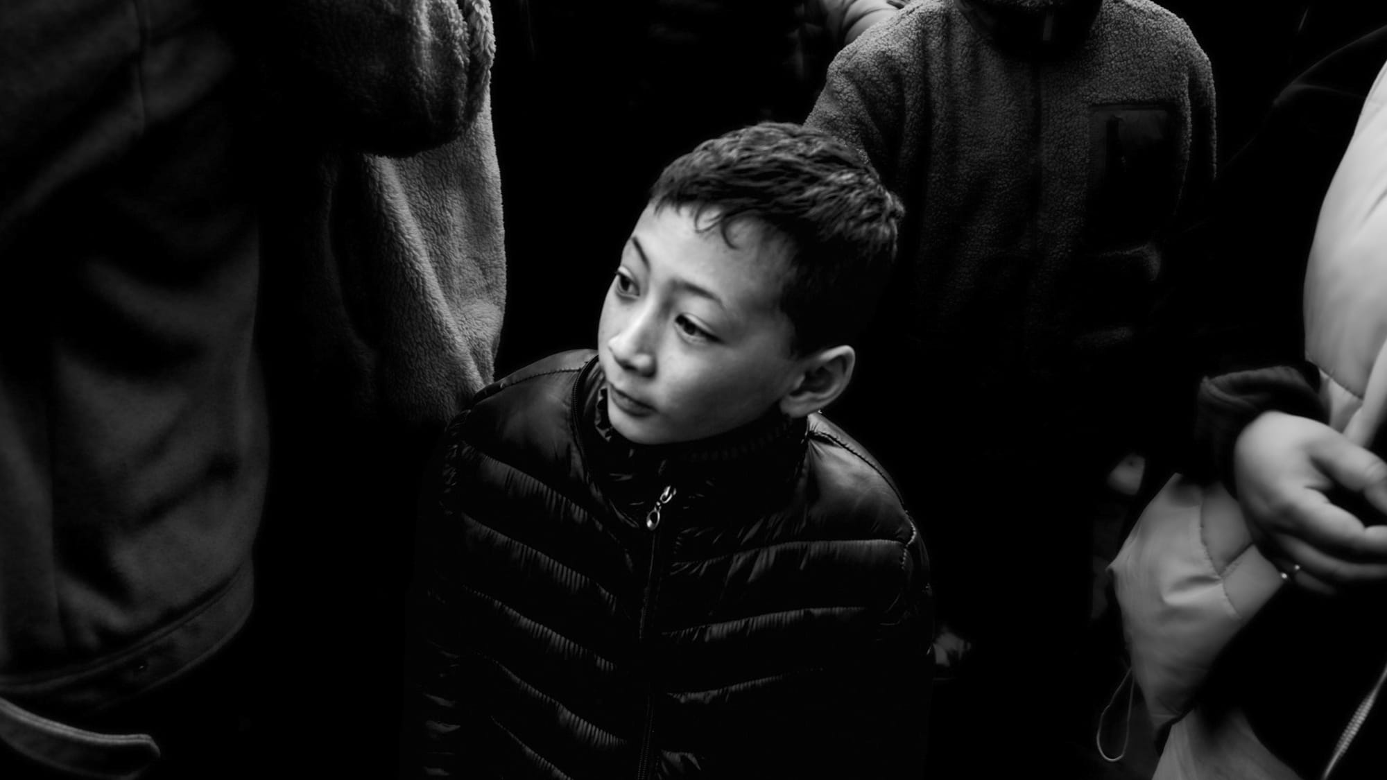 Black-and-white panoramic street portrait — a boy in a black puffer jacket, his face lit from above, gazes off to the side amid a dark press of adult bodies at the Leh festival.