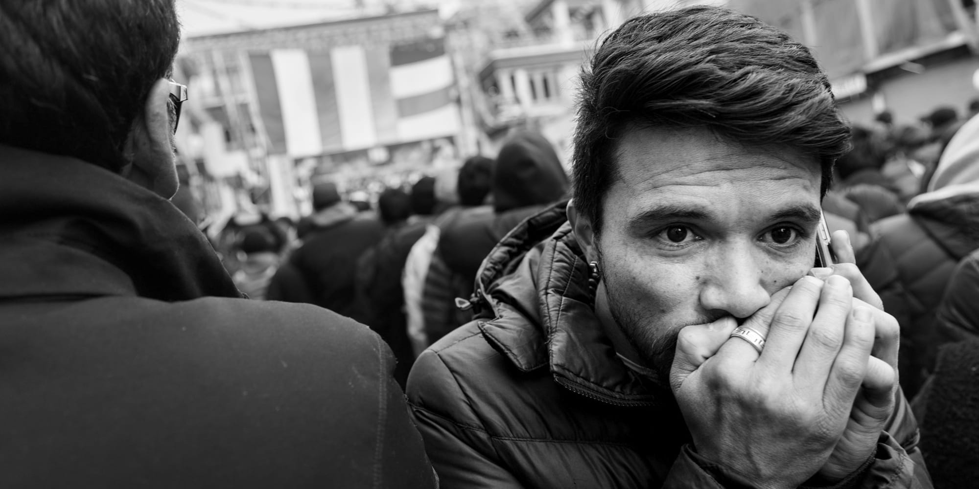 Black-and-white panoramic street photograph — a young man presses a phone to his lips, wide-eyed and waiting, as the Losar festival crowd and banners stretch behind him in Leh.