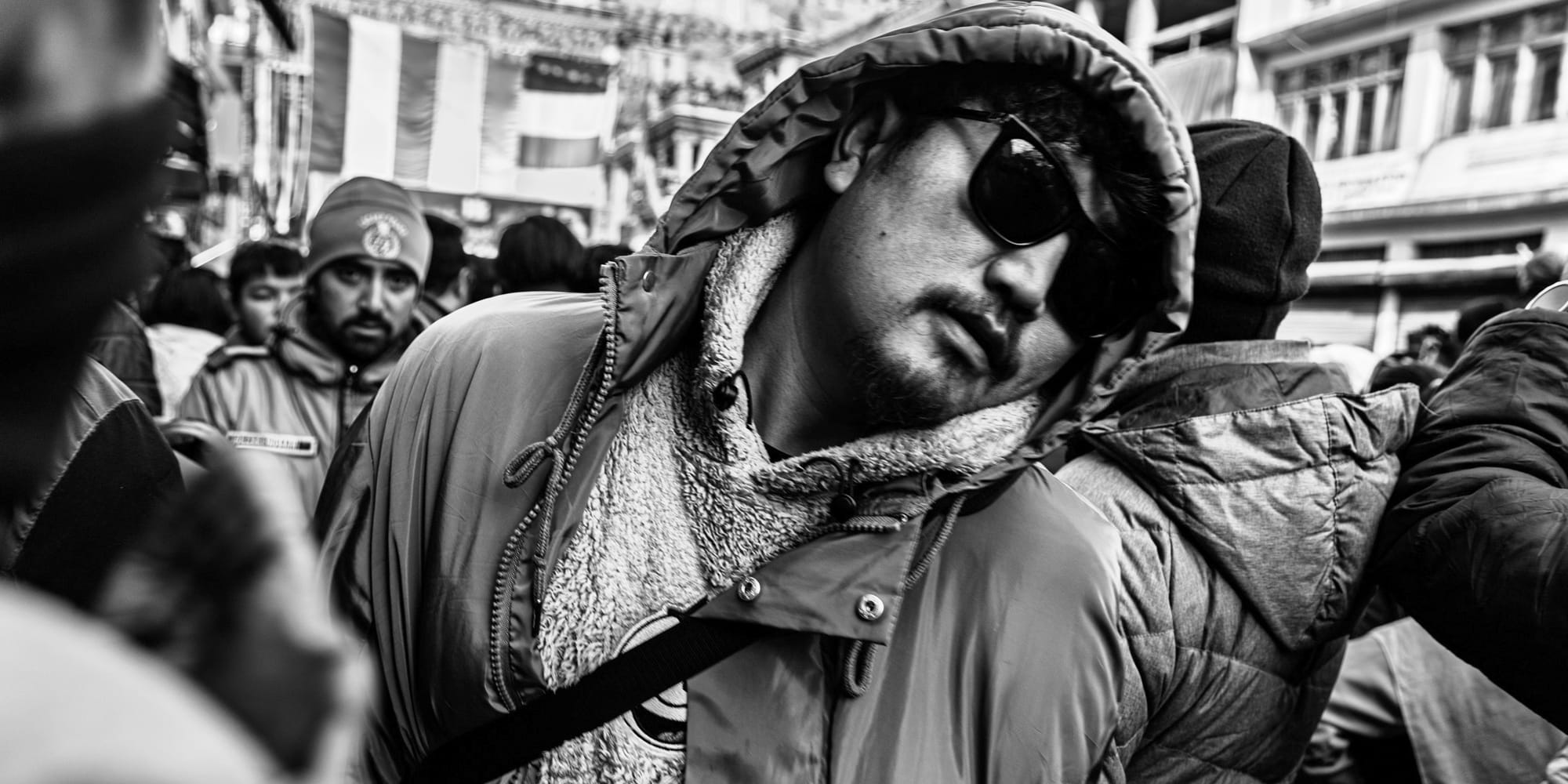 Black-and-white panoramic street photograph — a man in sunglasses and a fur-lined hood turns mid-stride with easy swagger through the packed festival street.