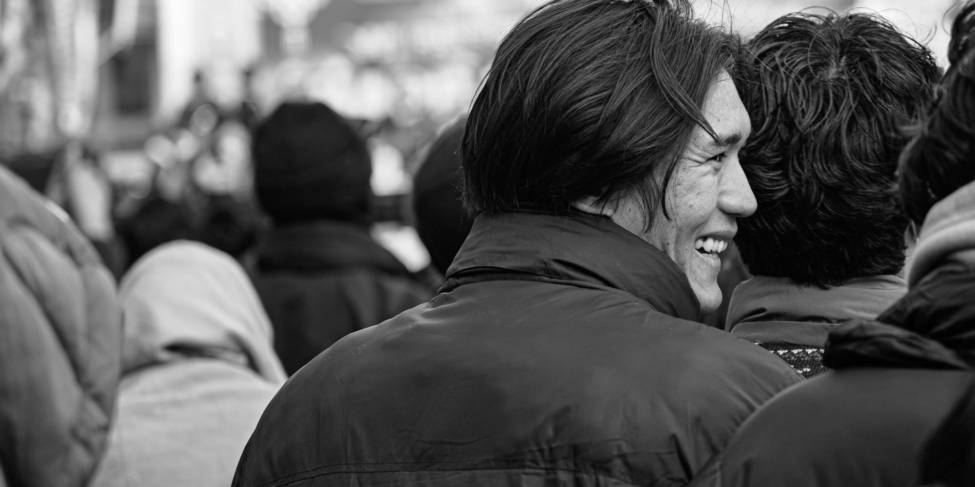Black-and-white panoramic street photograph — a young man laughs in profile, leaning cheek-to-cheek with a friend, the festival crowd dissolving into soft focus behind them.
