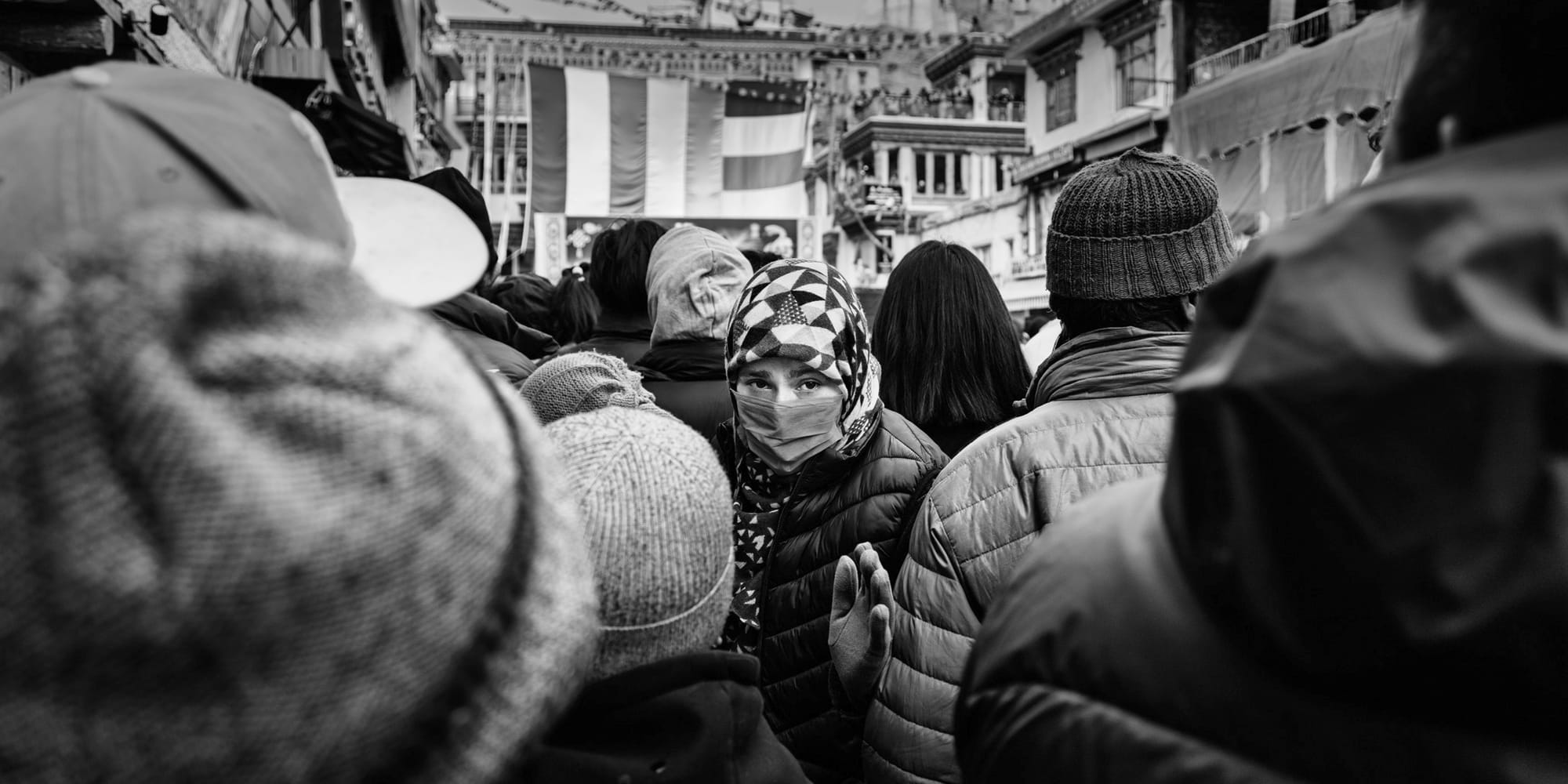 Black-and-white panoramic street photograph — a single masked face in a patterned cap meets the camera through a tunnel of hooded shoulders, festival banners rising over the Leh crowd.