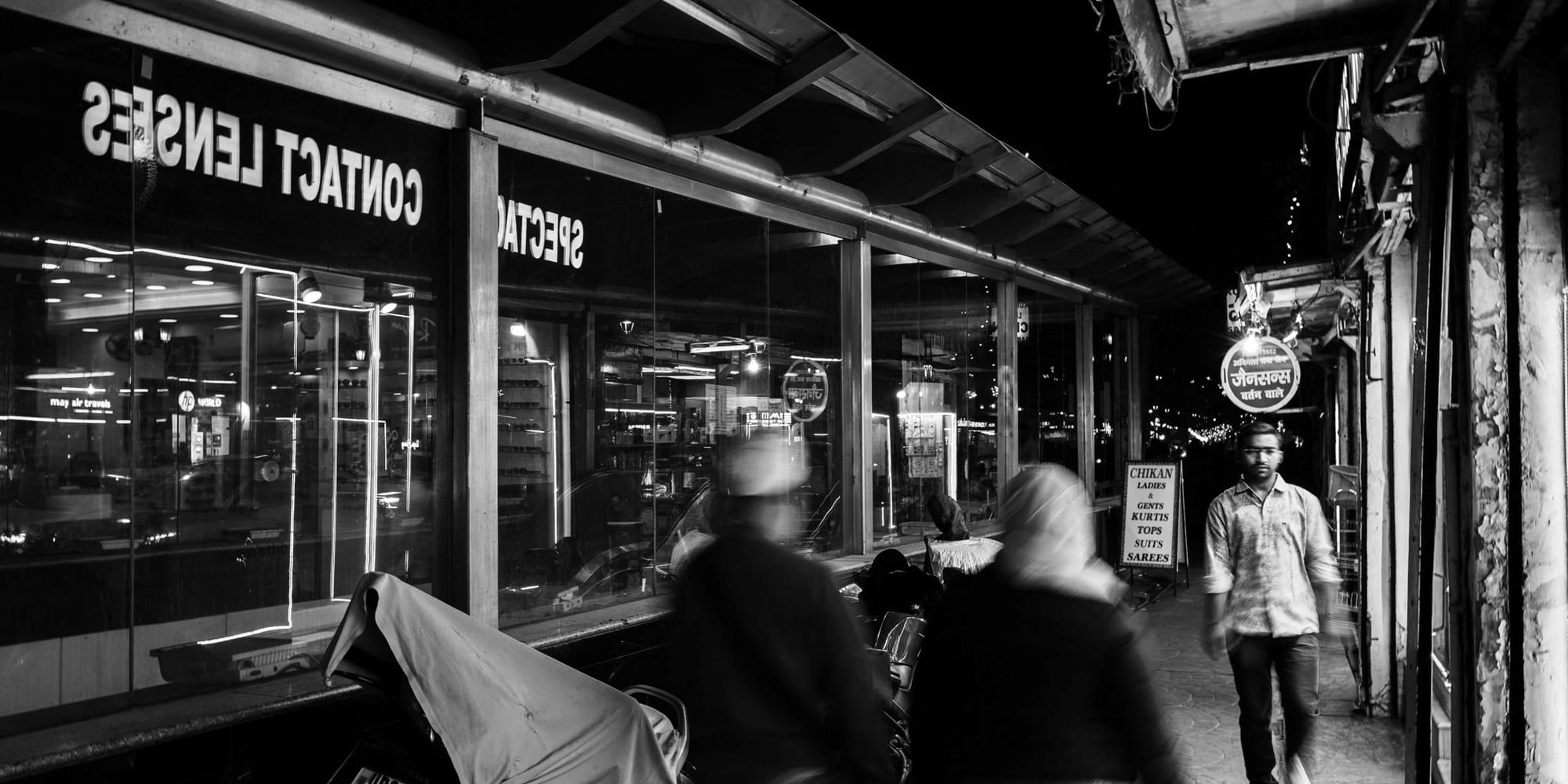 Monochrome long-exposure street scene — motion-blurred pedestrians ghost past a glass optician's storefront reading "Contact Lenses · Spectacles" as a lone man walks sharp in the distance, Hazratganj at night.