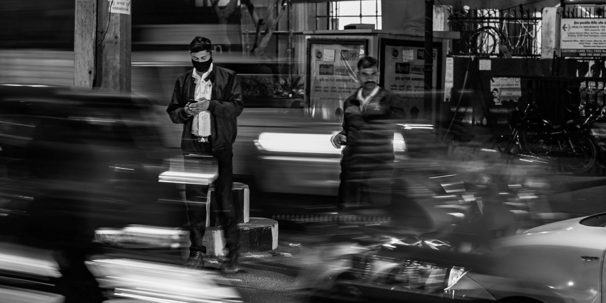 Slow-shutter black-and-white street photograph — a masked man stands still on a traffic island checking his phone while cars and motorbikes streak into light trails around him, the rush of Hazratganj at night.