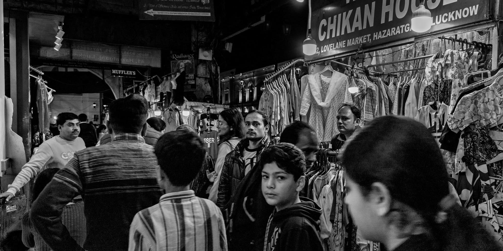 Monochrome street photograph in a crowded Hazratganj clothing market at night — shoppers move between racks of chikankari kurtas under a "Chikan House" sign, a young boy meeting the camera at center.