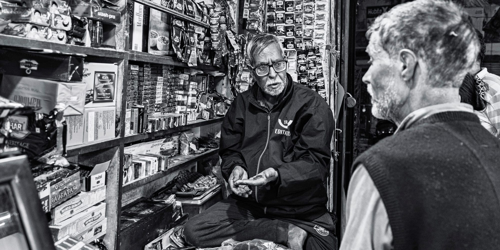 Black-and-white street photograph of a tightly packed Hazratganj cigarette-and-paan kiosk at night — an elderly shopkeeper counts coins in his palm as an older customer looks on, shelves crammed with packets behind them.