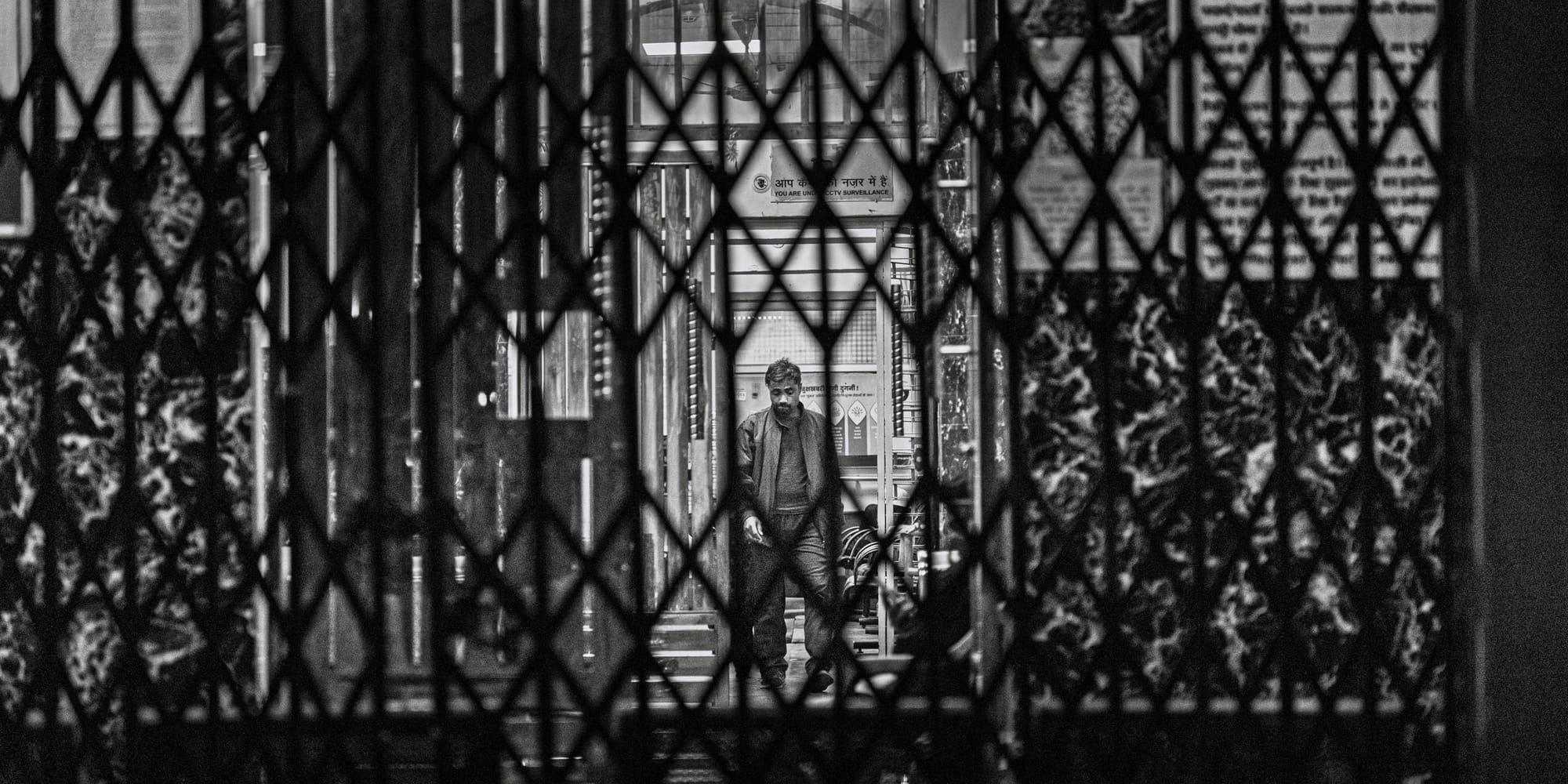Layered monochrome street frame shot through a diamond-pattern metal shutter — a man walks through a lit, tiled corridor beyond the defocused grille, Hazratganj at night.