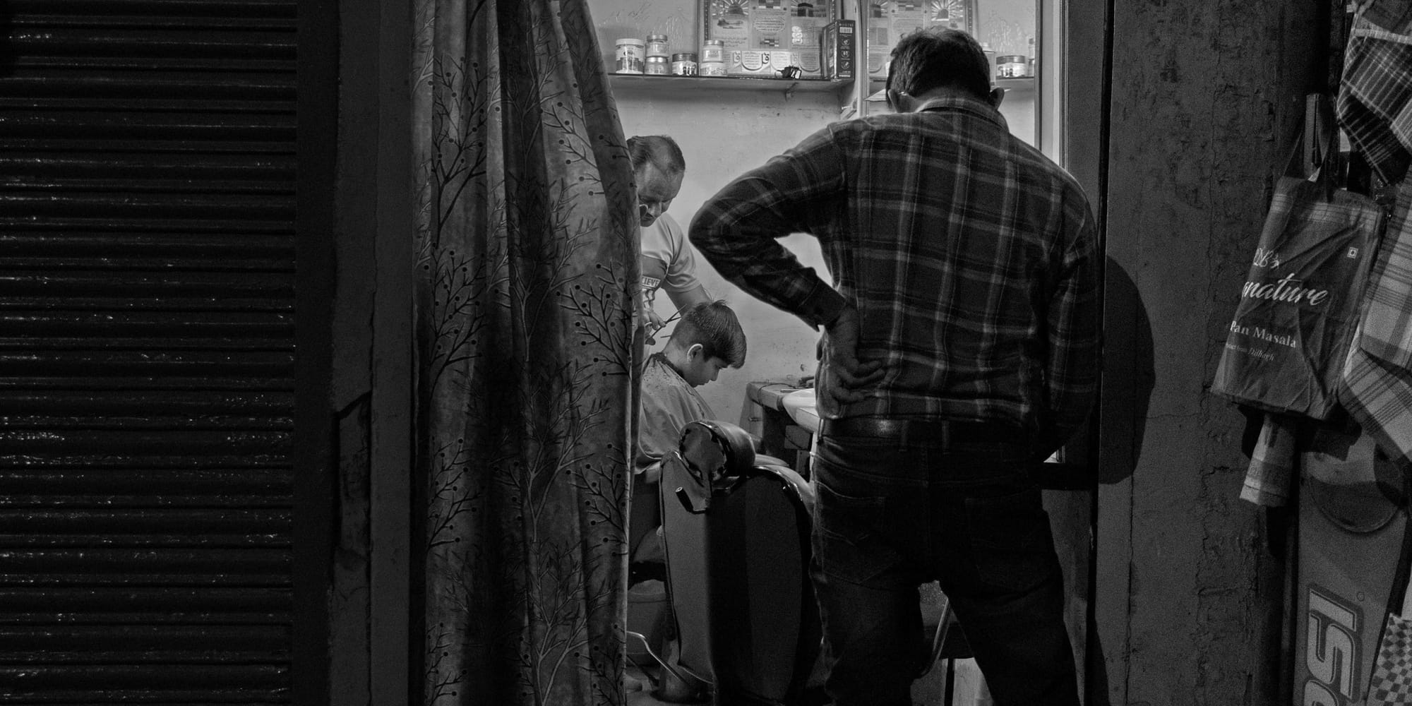 Candid black-and-white street photograph framed through a doorway — a barber trims a young boy's hair in a small Hazratganj salon at night, a waiting customer silhouetted in the foreground.
