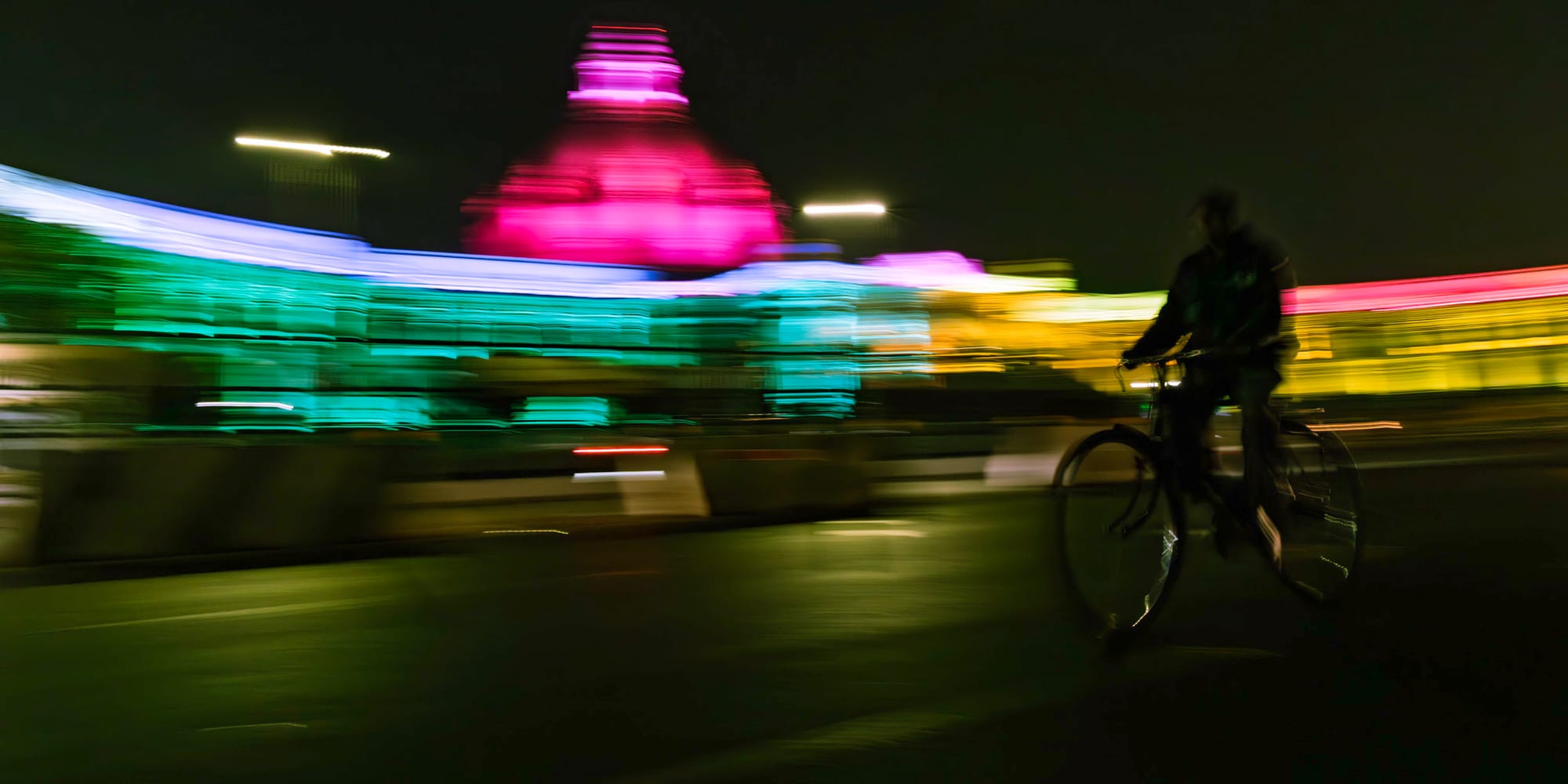 Panoramic slow-shutter panning photograph in color — a cyclist rides past the rainbow-floodlit dome of Hazratganj's assembly building at night, its facade dissolved into streaks of magenta, green and gold.