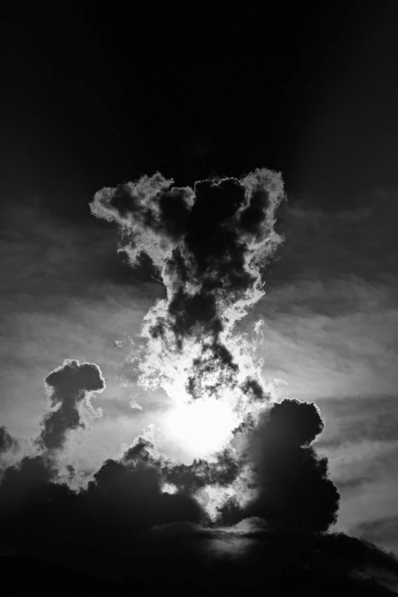  Black-and-white photograph — sunlight erupts through a towering, billowing cloud against an almost black sky.