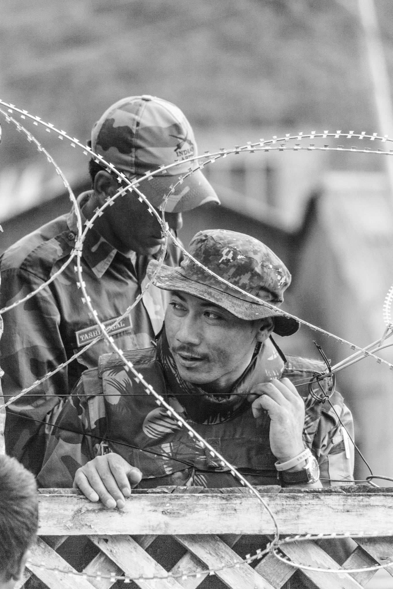 Black-and-white documentary photograph, two Indian Army soldiers in camouflage caps behind a coil of concertina razor wire, one leaning watchfully on a wooden barricade.