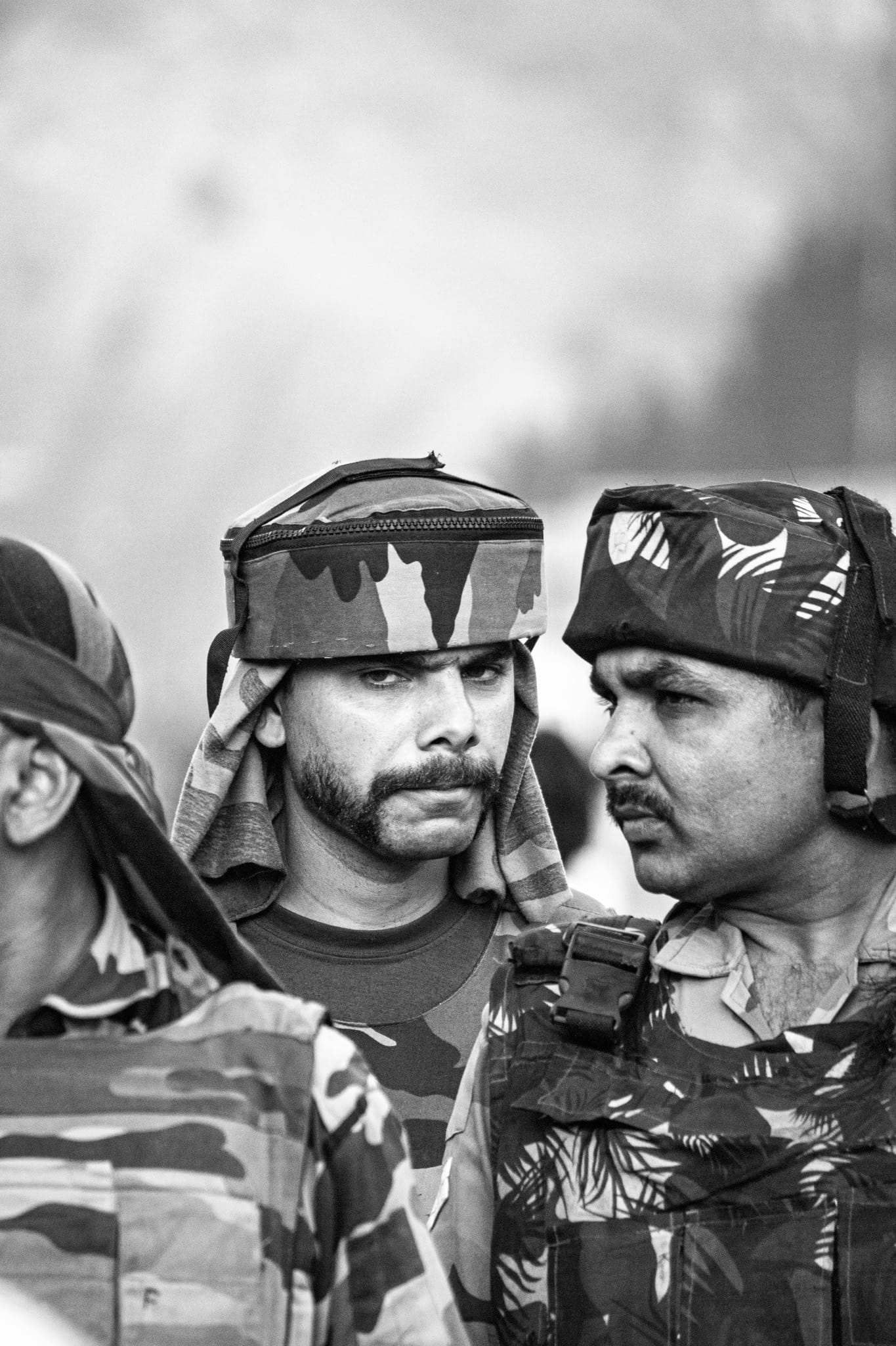 Black-and-white documentary portrait — three Indian soldiers in camouflage helmet covers and cloth head-wraps pressed shoulder to shoulder, faces set and watchful.