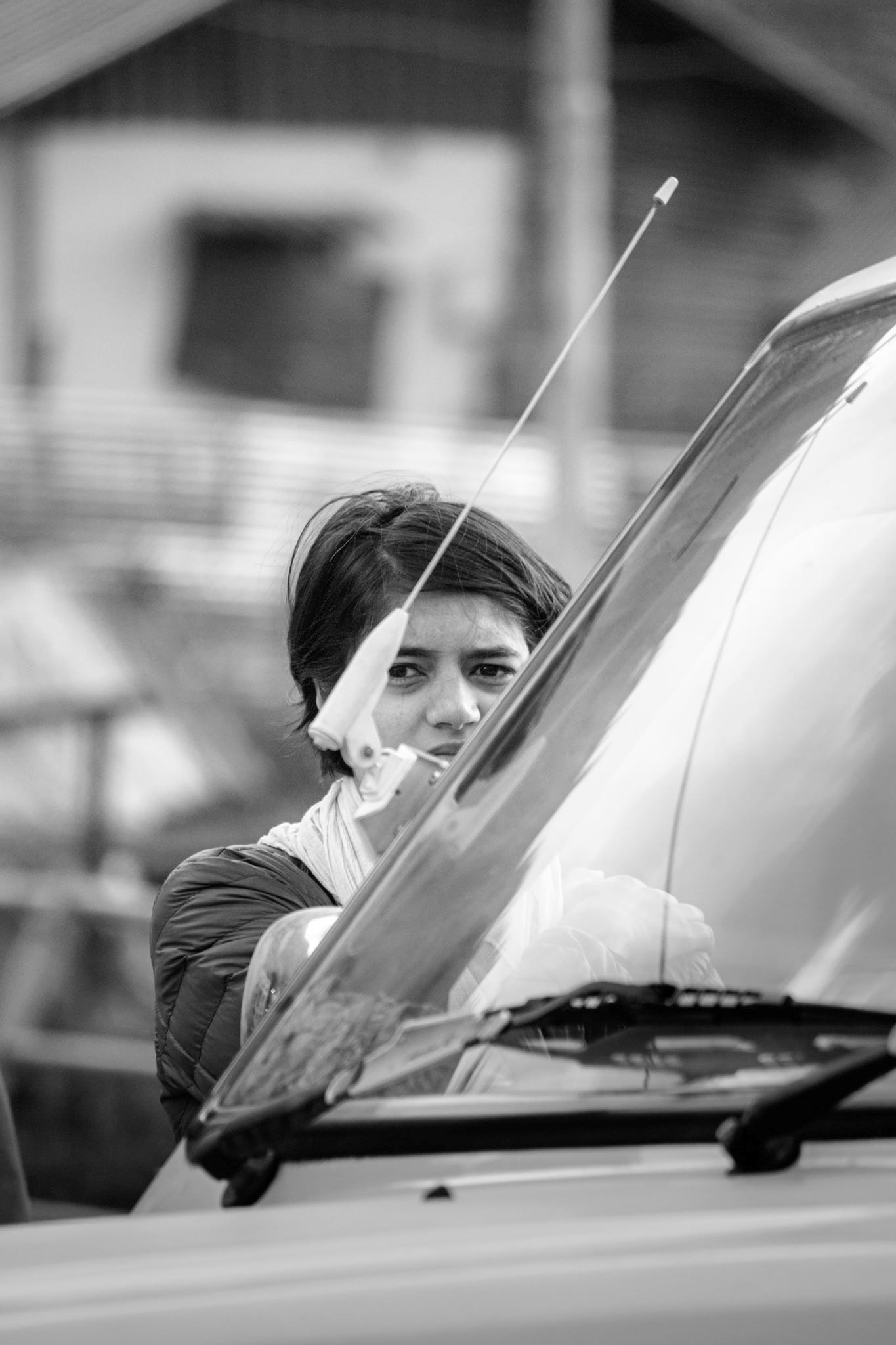  Black-and-white documentary photograph — a young woman in a dark jacket peers warily over a car's windshield, a thin radio antenna crossing her face.