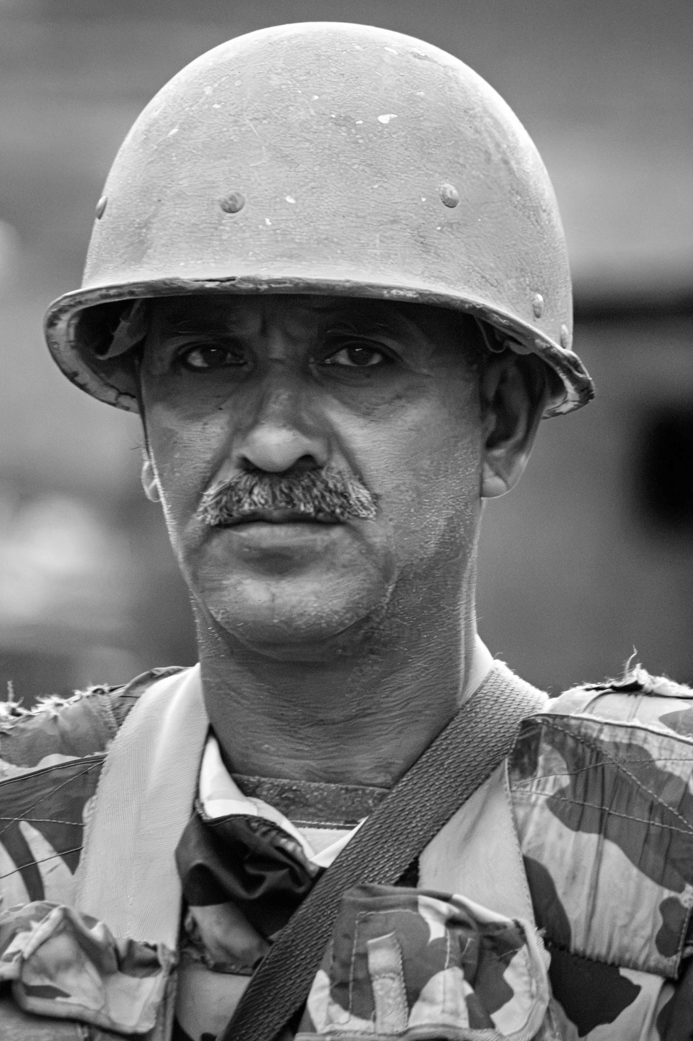 Black-and-white documentary portrait — a weathered older soldier in a battered steel helmet and camouflage webbing looks straight into the camera.