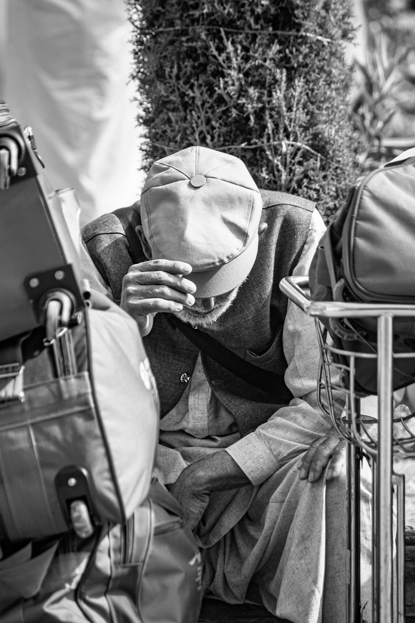Black-and-white documentary photograph — an elderly man in a flat cap and waistcoat bows his head into his hands, seated among suitcases and a luggage trolley.