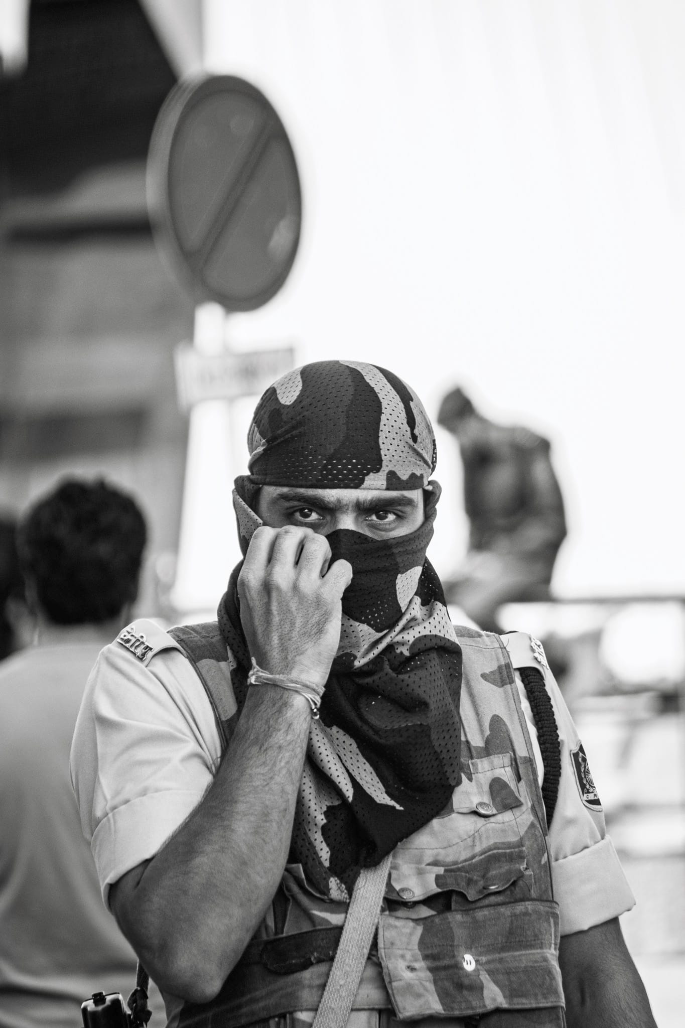 Black-and-white documentary photograph — a young paramilitary trooper draws a camouflage scarf up over his face until only his eyes show, a road sign and blurred figures behind.