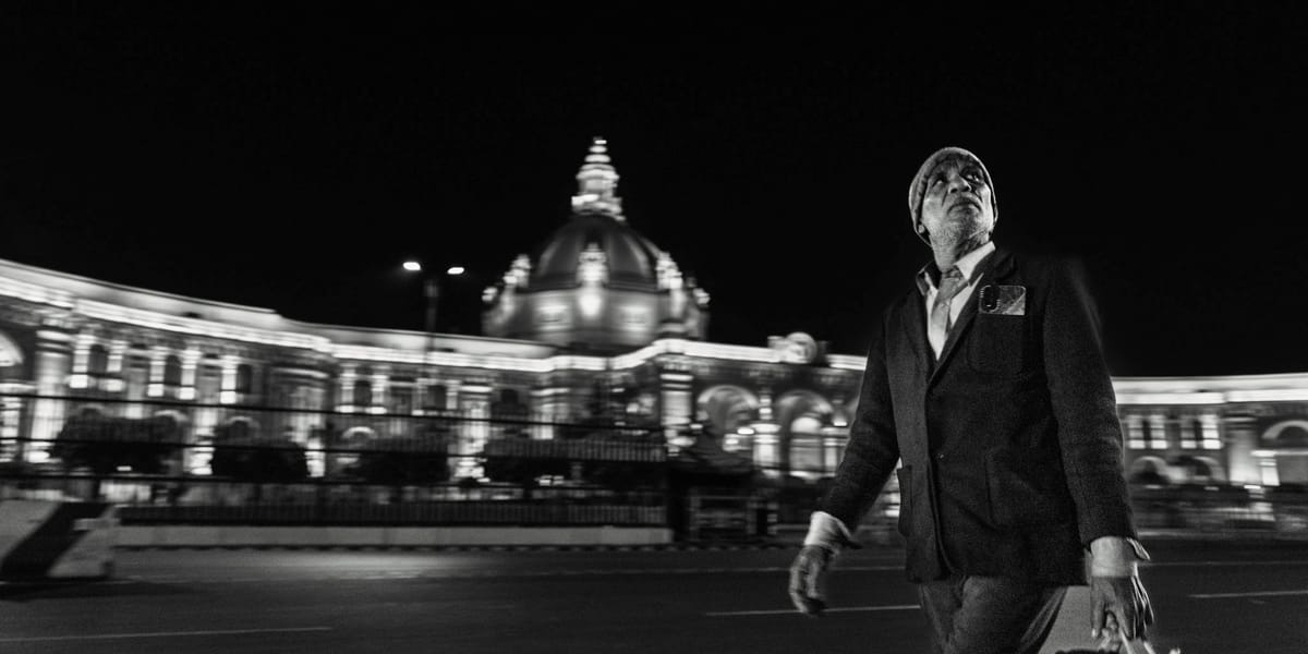 Black & white panoramic street photograph.a man in a flat cap and blazer strides past the floodlit Vidhan Sabha in Hazratganj at night, the architecture softened by slow-shutter pan blur.