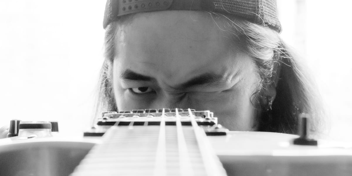 Black-and-white close-up — a long-haired musician in a mesh cap sights down the neck of an electric guitar, eyes narrowed over the strings as if taking aim.