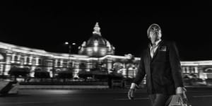 Black & white panoramic street photograph.a man in a flat cap and blazer strides past the floodlit Vidhan Sabha in Hazratganj at night, the architecture softened by slow-shutter pan blur.